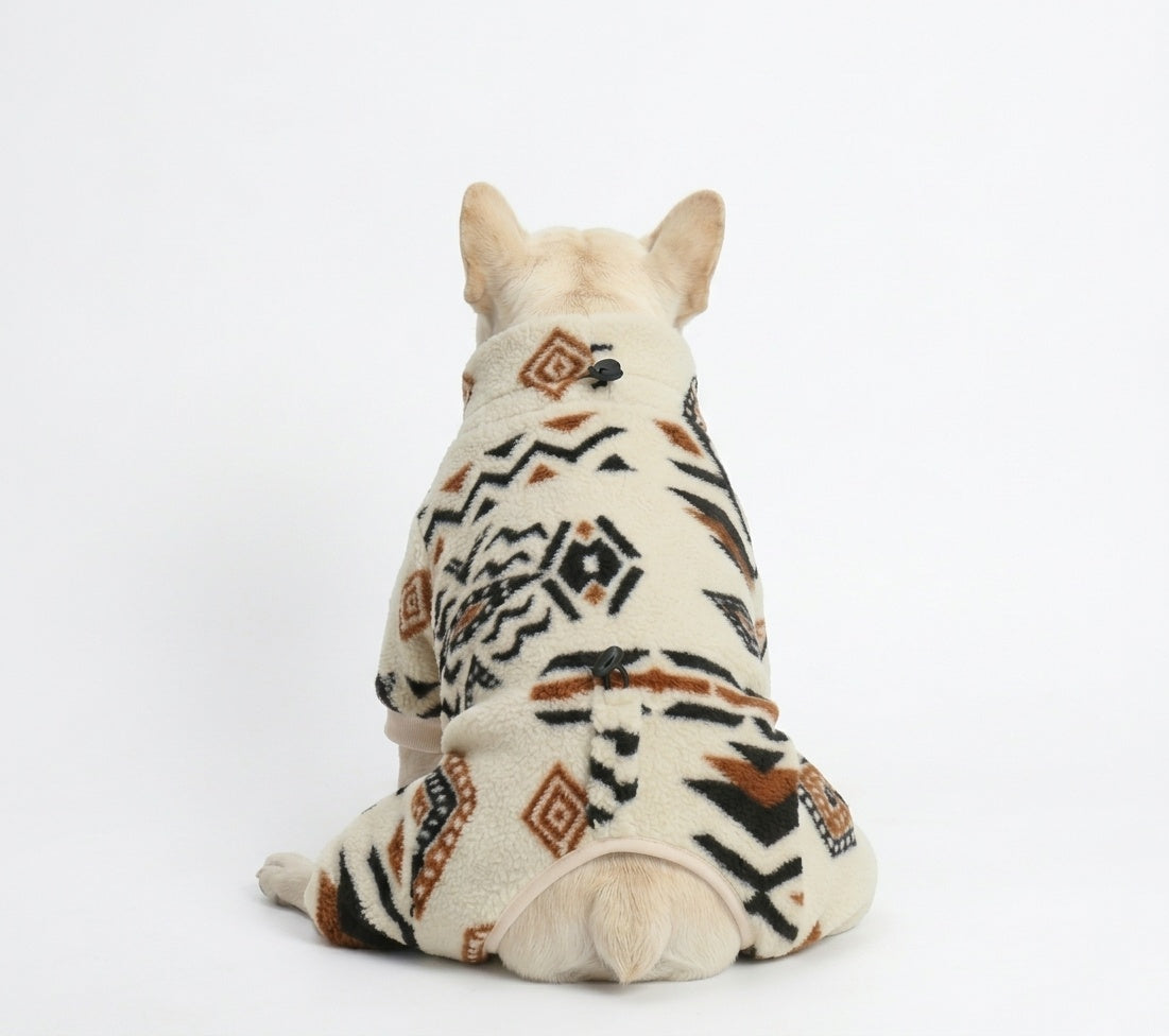 Rear view of a cream French Bulldog wearing a cream fleece four-legged onesie with black and brown tribal geometric patterns, featuring a neck toggle and tail opening on a white background.