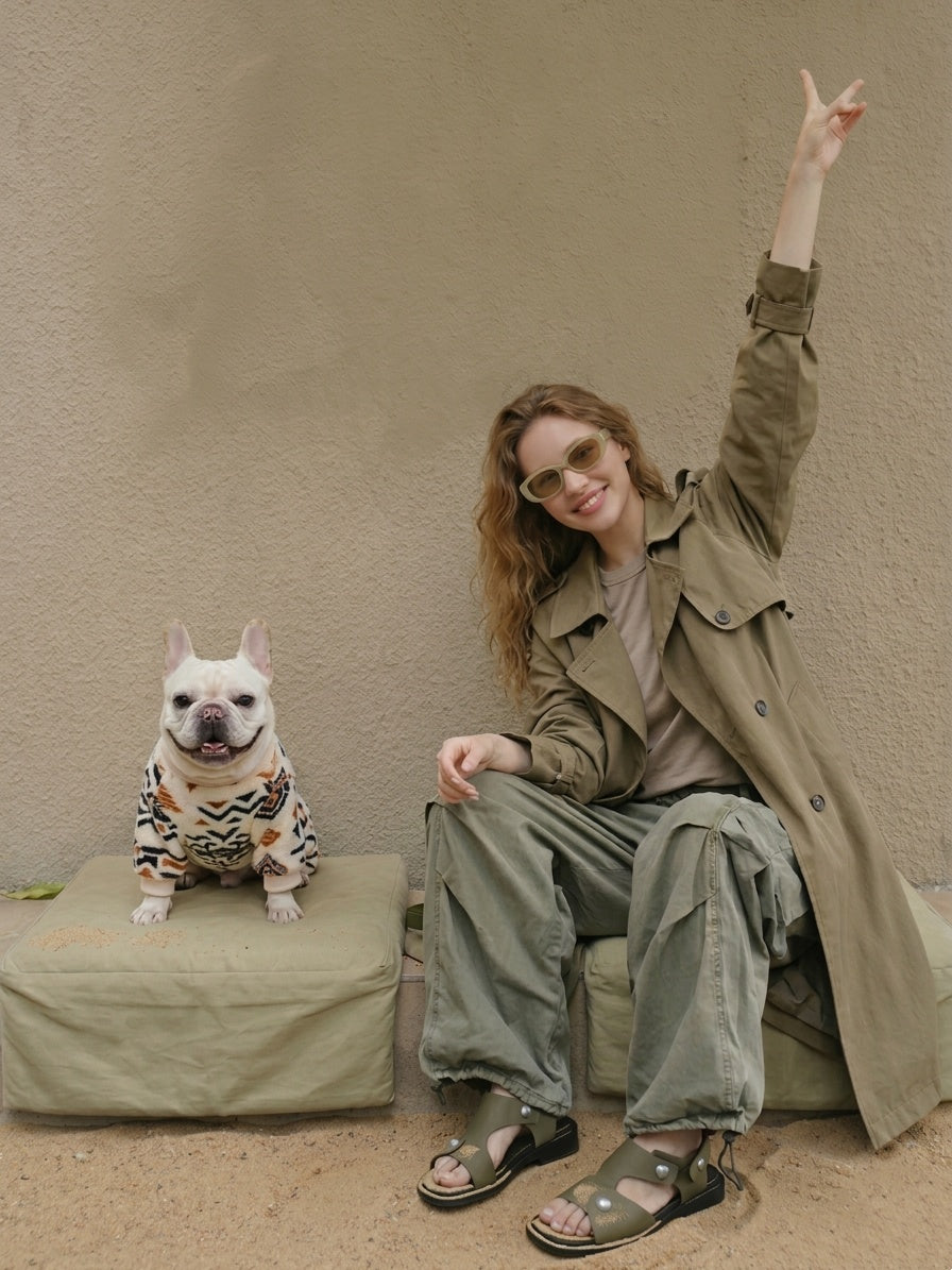 A 23-year-old Russian woman with a radiant smile, wearing a khaki trench coat and light khaki sunglasses, sitting beside a smiling cream-colored French Bulldog. The dog is wearing a bohemian tribal-patterned four-legged jumpsuit and is perched on a light beige cushion over desert sand. The background is a minimalist, solid-colored textured wall.